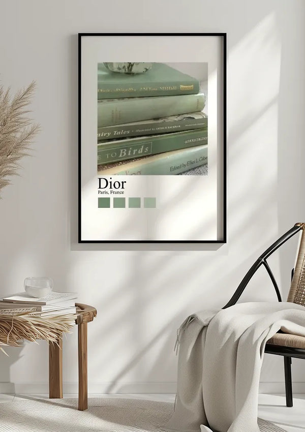 Een ingelijst CollageDepot Vintage Boeken Dior Parijs Schilderij met vintage boeken is op een witte muur gemonteerd. Hieronder staat een houten tafel met een keramieken voorwerp, naast een stoel met een witte deken eroverheen. Natuurlijk licht werpt schaduwen en accentueert de wanddecoratie.,Zwart