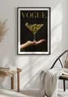 Een ingelijst Vogue-coverschilderij met de titel "VOGUE" met een groot martiniglas gevuld met groene olijven, vastgehouden door een hand met rode nagellak, hangt aan een witte muur. Daaronder staat een rieten stoel, gedrapeerd met een lichtgekleurde deken en een klein bijzettafeltje met boeken. Deze elegante wanddecoratie, het Vogue Cover Met Olijven Schilderij van CollageDepot, voegt verfijning toe aan elke ruimte.,Zwart