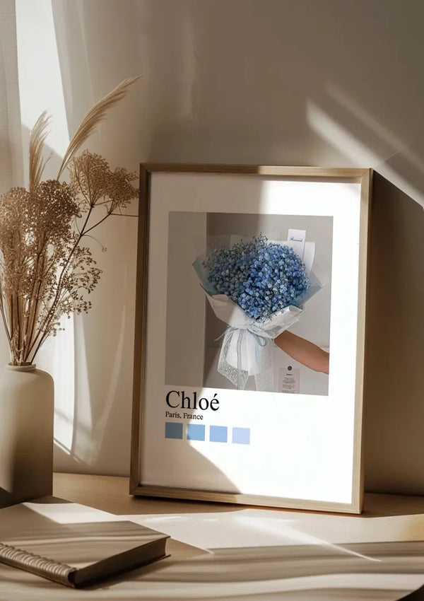 Een ingelijste foto van een hand die een boeket blauwe gipskruidbloemen vasthoudt, rust op een houten oppervlak. Ernaast staat een vaas met gedroogd, beige blad. Zonlicht werpt zachte schaduwen op de achterliggende muur, wat de charmante wanddecoratie extra benadrukt. De tekst op het frame luidt "Chloé Paris Schilderij" van CollageDepot.,Lichtbruin