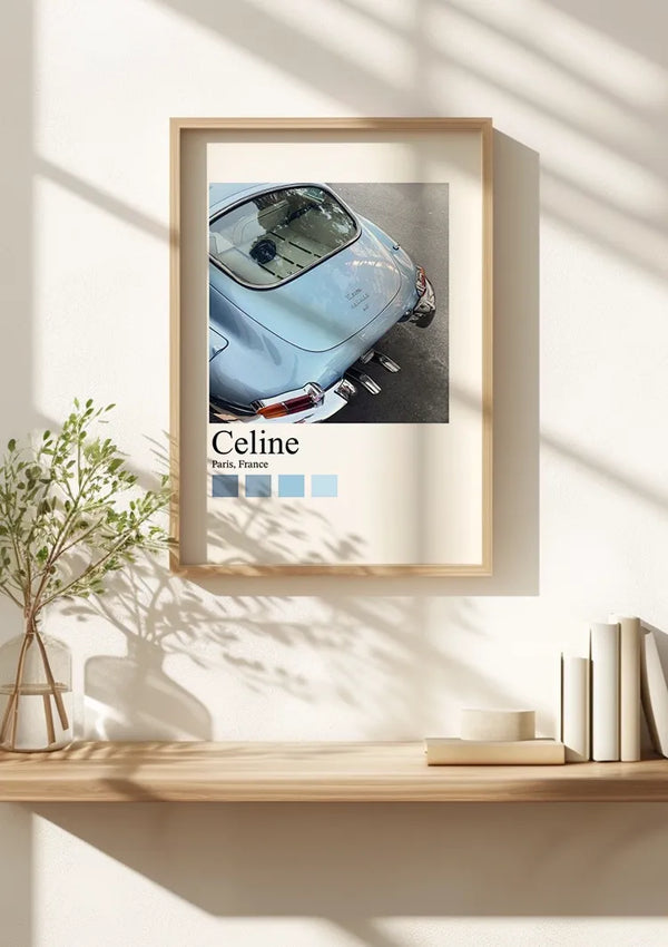 Een ingelijste poster hangt aan een witte muur boven een houten plank. Op de wanddecoratie staat een foto van de achterkant van een lichtblauwe vintage Porsche met het opschrift "Celine" met daaronder "Paris, France" geschreven. Op de plank staan diverse boeken en een potplant, alles beveiligd door een magnetisch ophangsysteem. Het kunstwerk, getiteld Vintage Porsche In Parijs Schilderij van CollageDepot, voegt charme toe aan de ruimte.,Lichtbruin