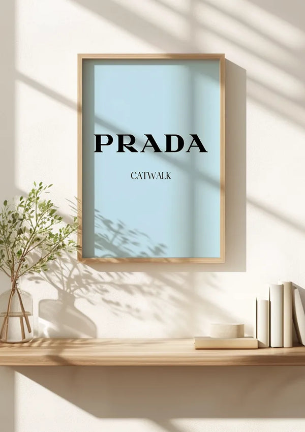 Een ingelijst CollageDepot Prada Catwalk Schilderij met de tekst "PRADA CATWALK" op een lichtblauwe achtergrond wordt als wanddecoratie op een muur getoond. Het staat op een houten plank naast een plantje in een glazen vaas en een stapel boeken aan de rechterkant, terwijl het zonlicht schaduwen werpt. Dit stuk maakt gebruik van een innovatief magnetisch ophangsysteem voor eenvoudige weergave.,Lichtbruin