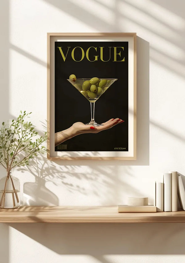 Een Vogue Cover Met Olijven Schilderij van CollageDepot met een opvallende hand die een martiniglas gevuld met groene druiven vasthoudt, hangt aan een lichtgekleurde muur met behulp van een magnetisch ophangsysteem. Onder deze elegante wanddecoratie bevindt zich een houten plank met een kleine plant en verschillende boeken, wat charme aan de ruimte toevoegt.,Lichtbruin