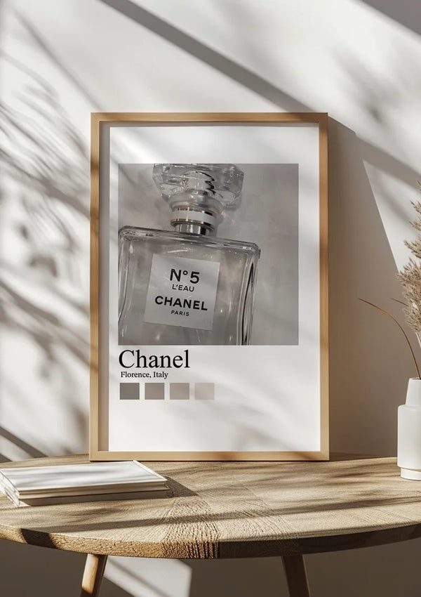 Op een houten tafel staat een ingelijste foto van een Parfumflesje Chanel N°5 Schilderij. Het frame bevat de tekst "CollageDepot Florence, Italië" onder de foto. Deze elegante wanddecoratie wordt aangevuld met een stapeltje boeken en een witte vaas met gedroogd gras.,Lichtbruin