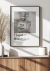 Een ingelijst Parfumflesje Chanel N°5 Schilderij van CollageDepot wordt aan een witte muur gehangen. De wanddecoratie wordt boven een houten kastje geplaatst met daarin een plantje en keramieken potjes. Natuurlijk licht en schaduwen van een nabijgelegen raam vallen op het toneel.,Zwart