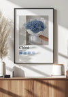 Een ingelijste foto van een blauw hortensiaboeket met de titel "Chloé Paris Schilderij" van CollageDepot hangt aan een witte muur, naadloos geïntegreerd in het decor als een prachtige wanddecoratie. Het frame, voorzien van een magnetisch ophangsysteem, zit boven een houten dressoir versierd met pampasgrasvaas, klein wit potplantje en boeken. Zonlicht werpt schaduwen op de muur.,Zwart