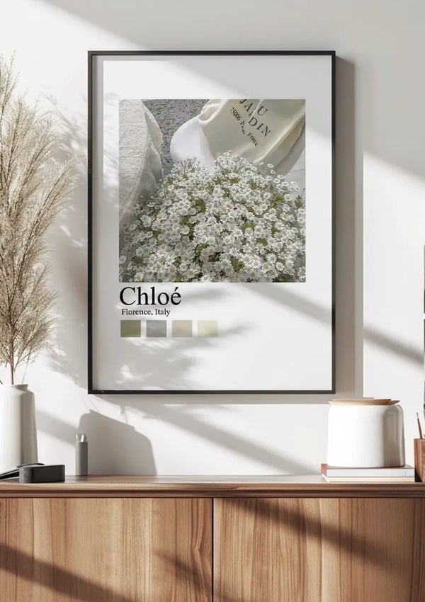 Een ingelijste Witte bloemen Cloé Parijs Schilderij van CollageDepot hangt aan een muur boven een houten dressoir, beveiligd door een magnetisch ophangsysteem. Op de poster staat een afbeelding van witte bloemen en een boek met de tekst “Chloé, Florence, Italy.” Op het dressoir staan decoratieve spullen waaronder potjes en een vaas met gedroogd gras.,Zwart