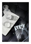 Een stijlvolle compositie met een CollageDepot ab 024 - zwart-wit glas half gevuld met water op een wit dienblad, naast een modetijdschrift, geplaatst op een zwartleren stoel onder natuurlijk licht.-