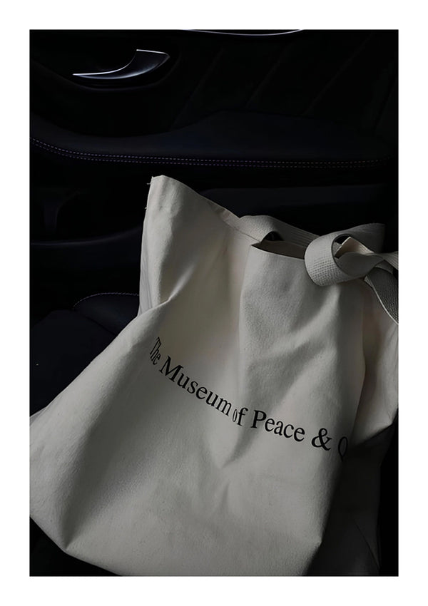Een witte draagtas met de tekst "The Museum of Peace & Art" in zwart gedrukt, rustend op een donkere autostoel, verlicht door natuurlijk licht.Productnaam: ab 047 - zwart-wit Merknaam: CollageDepot-