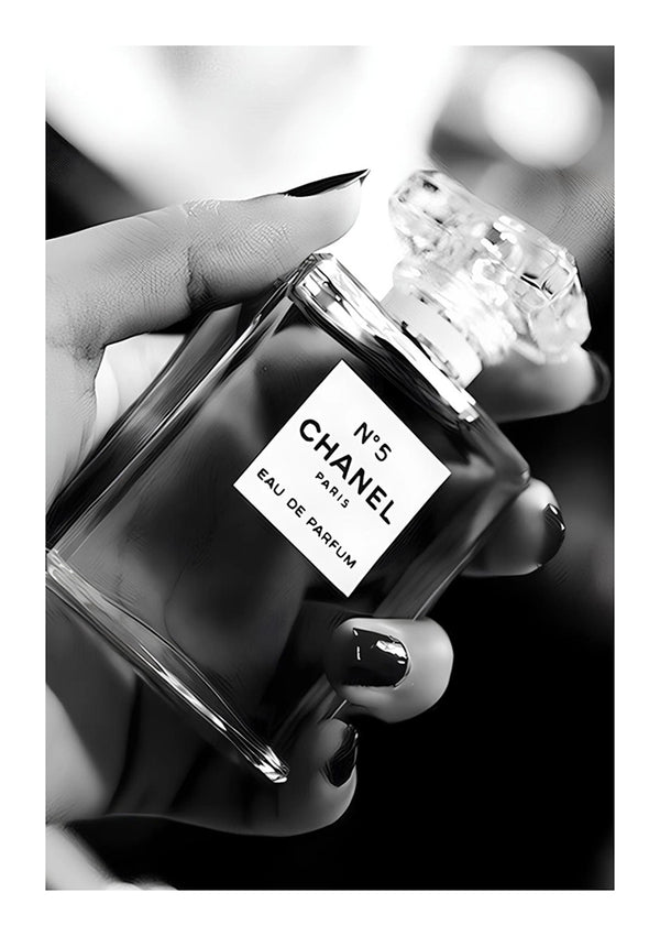 Een hand met verzorgde nagels houdt een fles Parfum In Hand Schilderij van CollageDepot vast. De heldere, rechthoekige fles met wit etiket en zwarte tekst steekt als een stijlvol accentstuk af tegen een onscherpe achtergrond, die doet denken aan wanddecoratie in een minimalistische kamer.-
