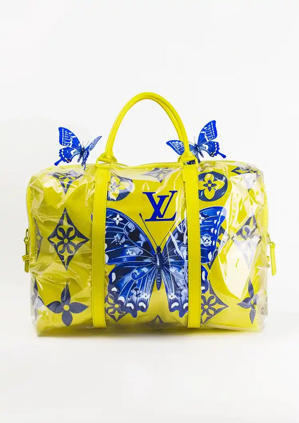Een groot, geel CollageDepot Louis Vuitton Gele Handtas Schilderij met blauwe vlindermotieven, waaronder het LV-logo en bloempatronen. De tas heeft gele handvatten, een doorzichtige plastic omhulling met extra blauwe vlinderaccenten en lijkt op een stijlvolle gele reistas, perfect voor elke reiziger.