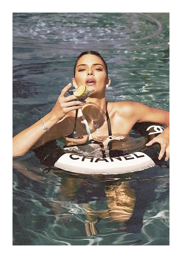 Een vrouw zit in een zwembad, met een martiniglas in haar hand. Ze draagt een zwempak en wordt ondersteund door een bb 003 - fashion float-ring van het merk CollageDepot. Ze ziet er ontspannen en glamoureus uit, gedeeltelijk ondergedompeld in water.-
