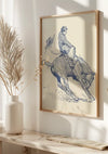 Een ingelijst Vintage Cowboy-schilderij van CollageDepot hangt aan een zonovergoten muur. Het toont een persoon die op een bokkend paard rijdt, terwijl een hoed wegvliegt. Op het oppervlak onder het kunstwerk wordt een vaas met gedroogd blad geplaatst, wat bijdraagt aan de moderne inrichting.,Lichtbruin