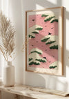 Aan een lichtgekleurde muur hangt een ingelijst schilderij Roze lucht van CollageDepot. Het kunstwerk heeft een roze achtergrond met groene en witte wolken en zwarte vogels tijdens de vlucht. Op de voorgrond staat een witte vaas met gedroogd gras op een witte plank. Zonlicht filtert door de scène, wat bijdraagt aan de serene Japanse stijl.