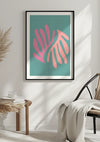 Aan een witte muur hangt een ingelijst abstract schilderij met roze bladachtige vormen op een blauwgroen achtergrond. Zonlicht werpt schaduwen in de kamer, die een rieten bijzettafeltje met gestapelde boeken en een glas bevat, en een houten stoel met een lichtgekleurde deken eroverheen. Het kunstwerk, bekend als het Tropical wonder Schilderij van CollageDepot, voegt een artistiek tintje toe aan de serene omgeving.