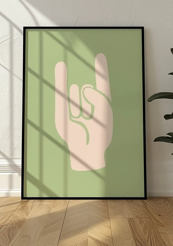 Een ingelijst Rock On Schilderij, leunend tegen een witte muur, toont een minimalistische hand die met twee opgeheven vingers een 'rock on'-gebaar maakt. De hand is geïllustreerd in lichte perzik op een gedempte groene achtergrond. Zonlicht en schaduw vallen over het schilderij en de houten vloer, wat deze moderne wanddecoratie van CollageDepot extra benadrukt.