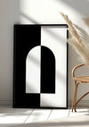 Een ingelijst minimalistisch Symmetrisch Zwart-Wit Schilderij van CollageDepot met geometrische vormen rust tegen een muur, voorzien van een magnetisch ophangsysteem. Vlakbij is een rieten stoel met geweven zitting en hoog siergras in een vaas zichtbaar. Zonlicht werpt schaduwen op de witte tegelvloer en muur, wat de serene sfeer versterkt.,Zwart
