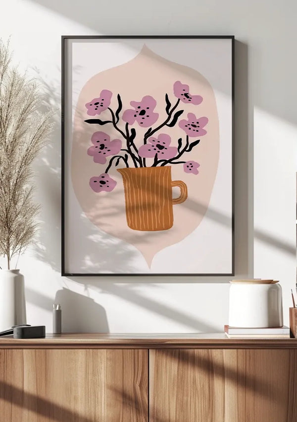 Een ingelijst abstract Kersenbloesem Schilderij van CollageDepot van een bruine mok met roze bloemen op takken hangt aan een witte muur boven een houten kast. De kast heeft een kleine potplant, een witte container en een bruin voorwerp. Zonlicht werpt schaduwen over de wanddecoratiescène.,Zwart