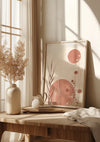 Een minimalistische interieurscène met een abstract Riet-schilderij van CollageDepot met roze cirkels en bloemvormen, geplaatst op een houten tafel. Op de tafel staan ook een witte keramische vaas, een kleine kan, gestapelde boeken en gedroogde bloemen. Beige gordijnen en zonlicht maken de setting compleet en versterken de zachte pasteltinten.,Lichtbruin