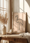Een minimalistisch CollageDepot Flamingo Schets Schilderij in een houten lijst rust tegen een muur op een houten tafel. Naast de wanddecoratie staan een potplant, een theepot, een stapel boeken en een doek. Het zonlicht stroomt door het raam naar binnen en werpt schaduwen.,Lichtbruin