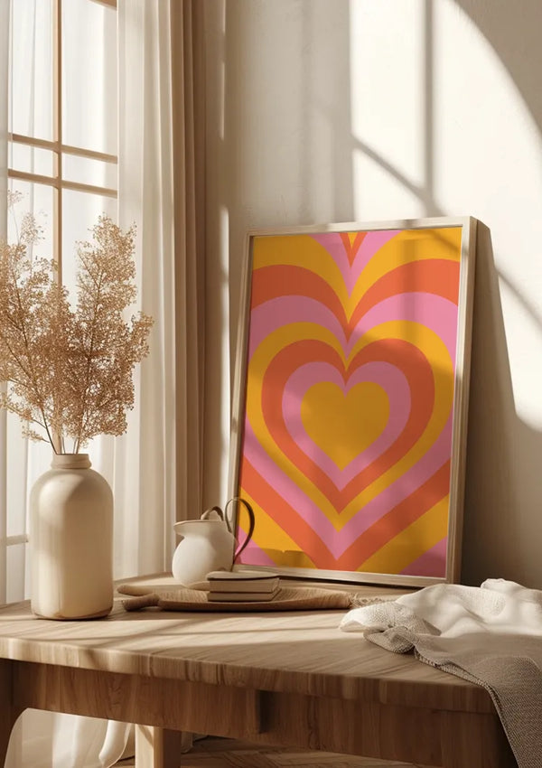 Een ingelijst CollageDepot Harten Patroon Schilderij met roze, oranje en gele patronen leunt tegen de muur op een houten tafel. Op de tafel staat ook een witte vaas met gedroogde bloemen, een stapel boeken, een theepot en een doek. Zonlicht filtert door een raam.,Lichtbruin