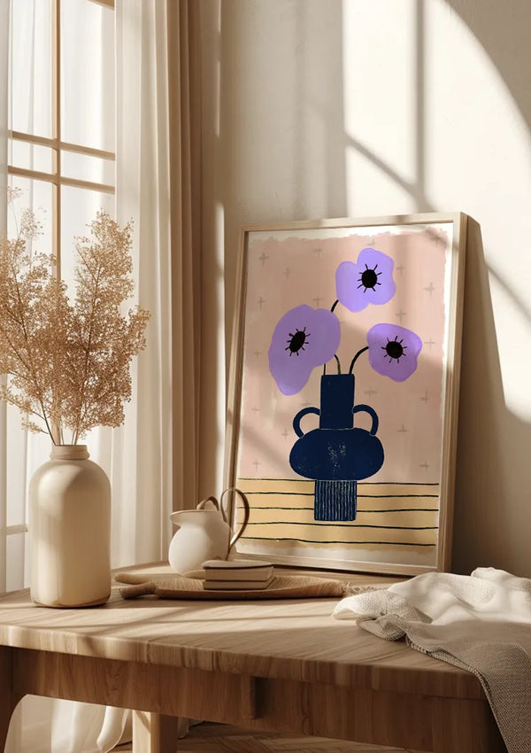 Op een houten tafel staat een ingelijste kunstprint met gestileerde bloemen in een vaas. Daarnaast zorgen een keramische vaas met gedroogde planten, een kleine witte kan en een boek voor extra charme. Zonlicht stroomt door het raam en werpt schaduwen op de Lavendelbloemen in Vaas Schilderij van CollageDepot en muur, waardoor deze elegante wanddecoratie nog beter tot zijn recht komt.,Lichtbruin