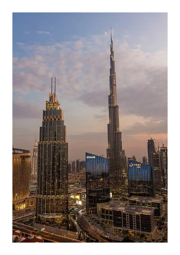 Een stadsgezicht bij zonsondergang bestaat uit verschillende wolkenkrabbers, waarvan de hoogste de Burj Khalifa is. Ernaast staat de Adres Boulevard-toren. De lucht is gedeeltelijk bewolkt en de stad wordt verlicht met lampen. Op de achtergrond zijn meerdere andere hoogbouw zichtbaar, zoals een groots Uitzicht Wolkenkrabbers En Busj Khalifa Schilderij van CollageDepot dat tot leven komt.-