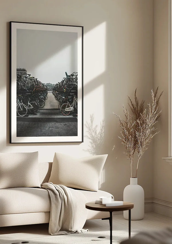 Een minimalistische woonkamer is voorzien van een beige bank met een lichte deken, een ronde houten tafel, een witte vaas met gedroogd pampasgras en een grote ingelijste foto aan de muur met een afbeelding van een drukke fietsenstalling. Het zonlicht stroomt van links naar binnen en versterkt de sfeer van Veel Fietsen Schilderij van CollageDepot.,Zwart