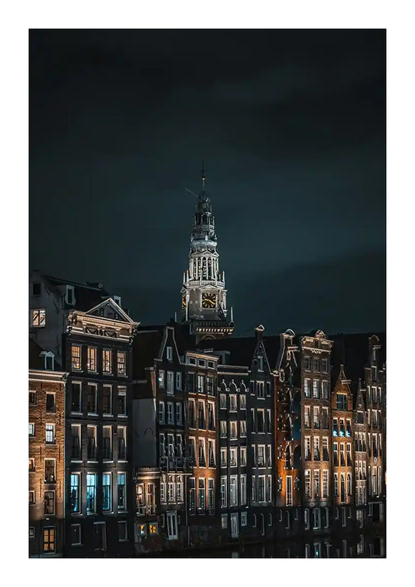 Een nachtscène met een rij verlichte gebouwen langs een gracht met een prominente klokkentoren op de achtergrond onder een donkere hemel. De gebouwen, die lijken op een Nachtelijk Tafereel Amsterdam Schilderij van CollageDepot, tonen traditionele Europese architectuur met opvallende, smalle gevels.-