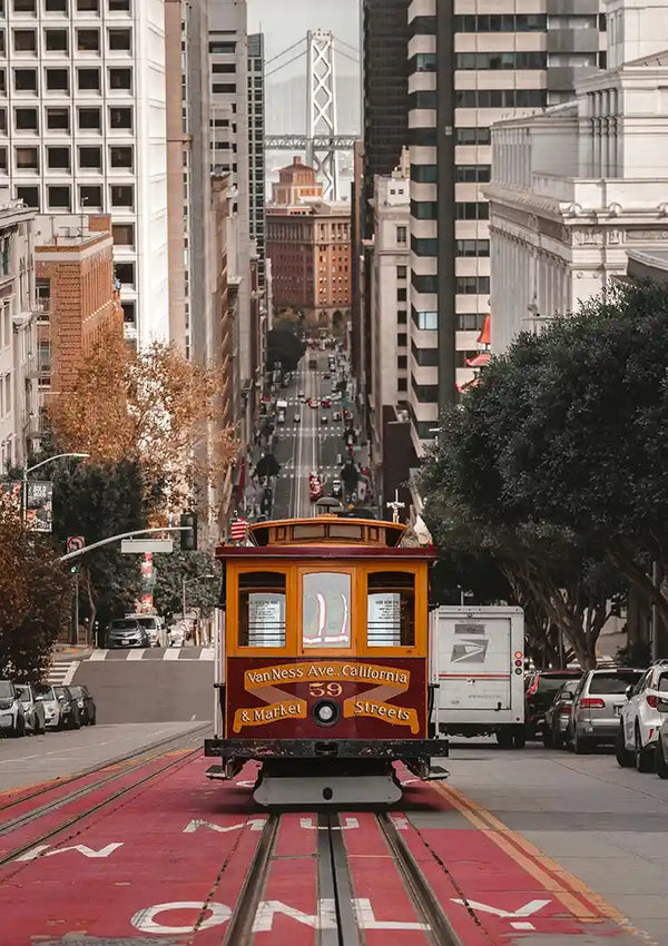 Een gele kabelbaan rijdt door een steile straat in San Francisco, omgeven door hoge gebouwen aan weerszijden. De straat, perfect voor een Tram Onderweg Schilderij van CollageDepot, is omzoomd met bomen en in de verte zie je een deel van de Bay Bridge. Aan beide zijden van de straat staan auto's geparkeerd.-