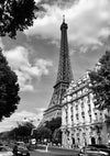 Zwart-witfoto van de Eiffeltoren in Parijs. Op de voorgrond zijn auto's en voetgangers zichtbaar op straat. De omgeving heeft klassieke Parijse architectuur en lommerrijke bomen, met daarboven een bewolkte hemel. Dit prachtige Elegante Gebouwen Met De Eiffeltoren Schilderij van CollageDepot dient als tijdloze wanddecoratie voor elke ruimte.-