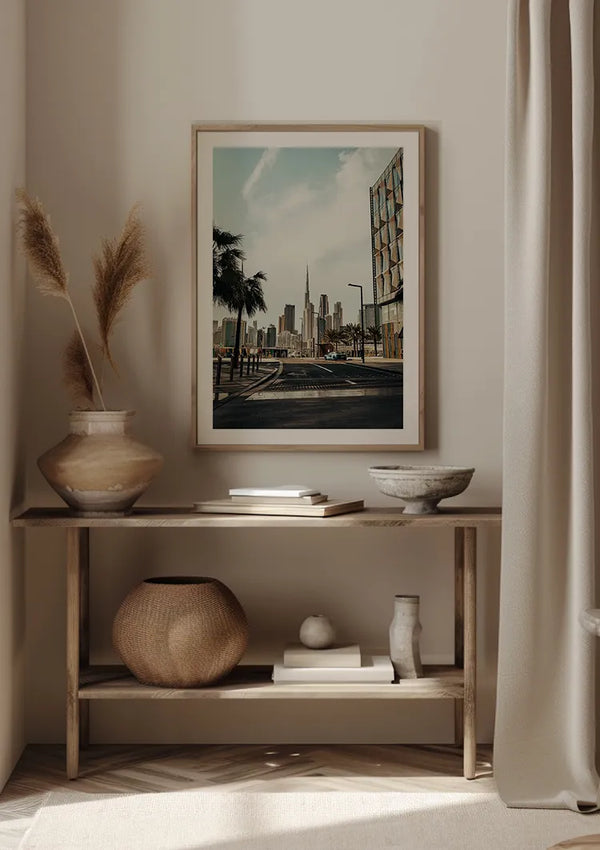 Een ingelijst Skyline Dubai schilderij van CollageDepot hangt aan een beige muur boven een houten consoletafel. Op de tafel staan verschillende decoratieve voorwerpen, waaronder een grote vaas met hoog gras, twee kommen, een potje en een geweven mand op de onderste plank.