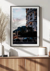 Een Hotel de Paris by night schilderij van CollageDepot is als stijlvolle wanddecoratie op een witte muur gemonteerd. Het beeld wordt boven een houten kast geplaatst die versierd is met decoratieve voorwerpen, waaronder een hoge vaas met gedroogd pampagras en een stapel boeken.