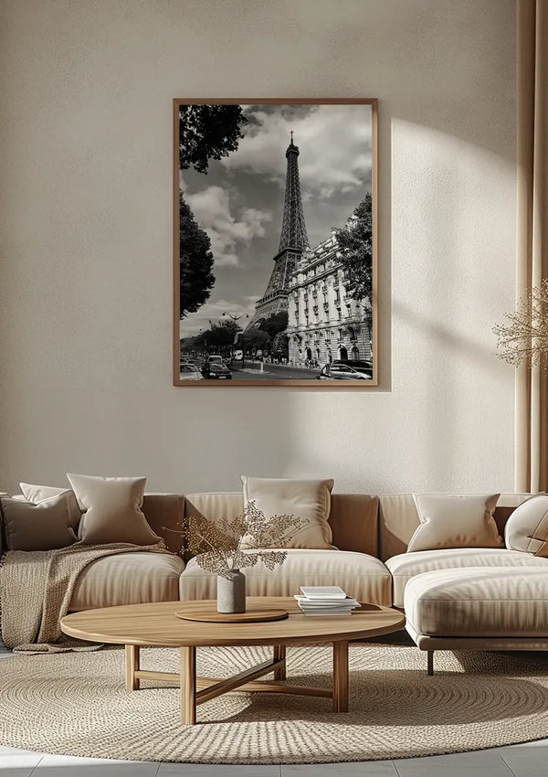 Een woonkamer met een beige kleurenschema is voorzien van een groot CollageDepot Elegante Gebouwen Met De Eiffeltoren Schilderij aan de muur boven een beige bank. In het midden staat een ronde houten salontafel, versierd met kleine decoratieve items.,Lichtbruin