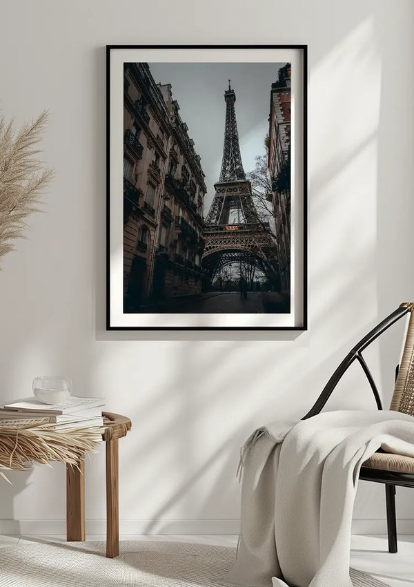 Een ingelijst Smalle Straat Eiffeltoren Schilderij van CollageDepot hangt aan een lichtgekleurde muur met behulp van een magnetisch ophangsysteem. Daaronder staat een geweven kruk met een boek en een kom, naast een stoel met beige kussens en een witte deken. Pampasgras siert de hoek van de kamer.,Zwart