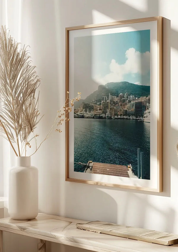 Een schilderij met uitzicht op Monaco van CollageDepot als middelpunt van deze wanddecoratie toont een haven met aangemeerde boten en gebouwen op een heuvel onder een bewolkte hemel. Op de voorgrond staat een witte vaas met gedroogd blad op een richel die baadt in natuurlijk licht, beveiligd door een innovatief magnetisch ophangsysteem.