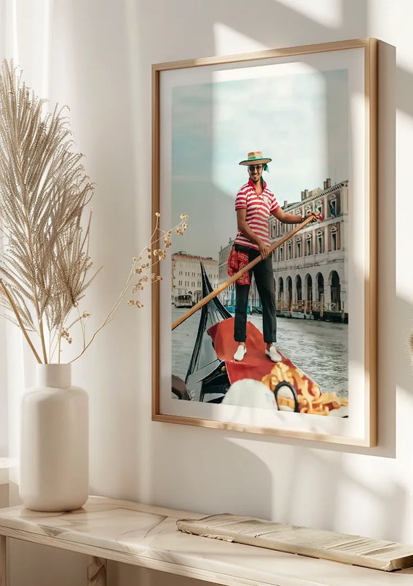 Een ingelijst Gondelier Venetië Schilderij van CollageDepot op een zonovergoten muur toont een gondelier in een gestreept overhemd en strohoed die een gondel roeit. De achtergrond bestaat uit Venetiaanse architectuur en water, wat doet denken aan een kanaaltafereel dat typisch is voor Venetië, Italië. In de buurt voegt een vaas met gedroogde planten charme toe aan deze elegante wanddecoratie.,Lichtbruin