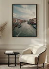 Een ingelijst Canal Grande Schilderij van CollageDepot is gemonteerd op een witte muur boven een crèmekleurige fauteuil met een bijpassende vloerlamp aan de zijkant. Links van de stoel staat een klein zwart bijzettafeltje met een stapel boeken. De kamer heeft een minimalistische, moderne inrichting, versterkt door deze elegante wanddecoratie.,Zwart