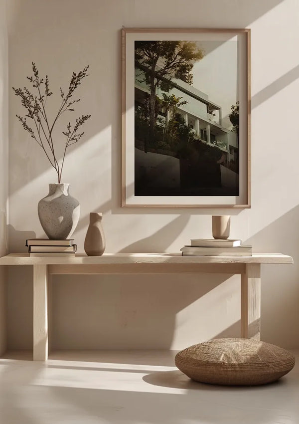 Een minimalistische kamer met een houten bank met twee vazen, boeken en een ingelijste foto van moderne architectuur. Op de grond ligt een rond geweven kussen. Zonlicht filtert er doorheen en werpt schaduwen. In een van de vazen staat een takje gedroogd blad. Aan de wand is een schilderij van Villa Marbella opgehangen met een magnetisch ophangsysteem van CollageDepot.
