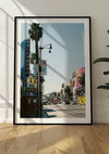 Een ingelijste foto van een drukke straat in Hollywood, gemonteerd op een lichtgekleurde muur met zichtbare schaduwen. De straat is omzoomd met palmbomen en gebouwen met kleurrijke advertenties en een verticaal 'Hollywood'-bord. Dit Hollywood Boulevard Schilderij van CollageDepot wordt veilig op zijn plaats gehouden met behulp van een magnetisch ophangsysteem.,Zwart