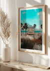 Een ingelijst CollageDepot Badmeestertoren Op Het Strand Schilderij dat aan een witte muur hangt, toont een strandtafereel met een badmeestertoren, palmbomen en zonnebadende mensen. Het tafereel is helder en helder, met een turquoise lucht. Onder de foto staat een kleine potplant op een witte tafel.,Lichtbruin