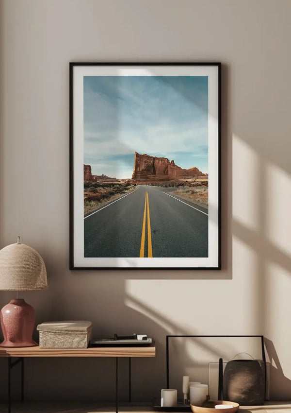 Aan een beige muur hangt een ingelijste foto van een woestijnweg met een gele scheidingslijn, wat bijdraagt aan de elegante wanddecoratie. Hieronder staat een houten plank met een lamp, boeken en decoratieve voorwerpen. Natuurlijk licht werpt schaduwen over de muur en de plank. Deze scène toont het Desert Road-schilderij van CollageDepot.
