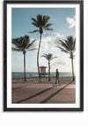 Een ingelijste foto, perfect als wanddecoratie, toont een strandtafereel met vier hoge palmbomen en een persoon die over een verhard pad loopt. De oceaan is zichtbaar op de achtergrond onder een gedeeltelijk bewolkte hemel. Bij het water staat een badmeestertoren, met schaduwen van de bomen en de persoon die zich op de grond uitstrekt. Het product "Het Strand Met De Palmbomen Schilderij" van CollageDepot legt dit pittoreske moment prachtig vast.,Zwart-Met,Lichtbruin-Met,showOne,Met