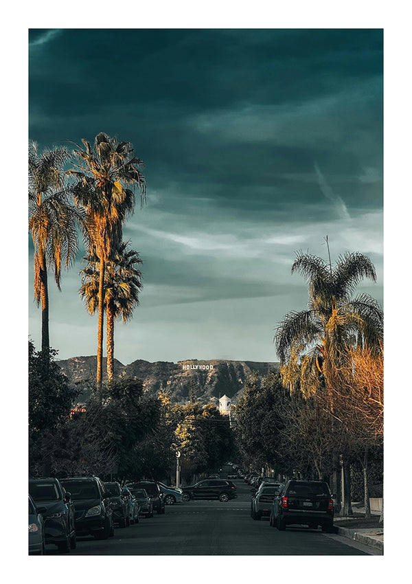 Een straat met geparkeerde auto's leidt naar het Hollywood-bord, zichtbaar op de heuvel in de verte. Hoge palmbomen flankeren de straat en de lucht is gedeeltelijk bewolkt met donkere en lichte plekken. De scène baadt in een warm, gouden licht, alsof een CollageDepot Hollywood Bord In De Verte Schilderij een perfect moment vastlegt.-