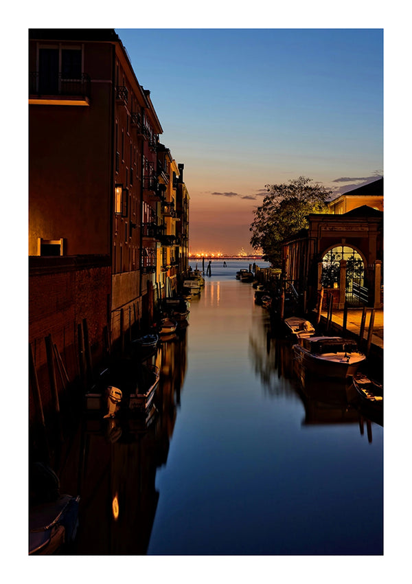 Bij zonsondergang wordt een smal kanaal met gebouwen en kleine boten afgebeeld. De lucht verandert van blauw naar oranje terwijl de zon ondergaat aan de horizon. Het water reflecteert het vervagende licht, waardoor een serene en rustige sfeer ontstaat, perfect voor een Zonsondergang In Venetië Schilderij met een magnetisch ophangsysteem van CollageDepot.-