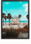 Een ingelijst Badmeestertoren Op Het Strand Schilderij van CollageDepot toont een strandtafereel met een houten badmeesterhut op palen, omgeven door palmbomen. Mensen ontspannen op het zand onder een strakblauwe lucht, met duingras op de voorgrond en de oceaan op de achtergrond. Deze prachtige wanddecoratie is voorzien van een magnetisch ophangsysteem voor eenvoudige presentatie.,Zwart-Zonder,Lichtbruin-Zonder,showOne,Zonder