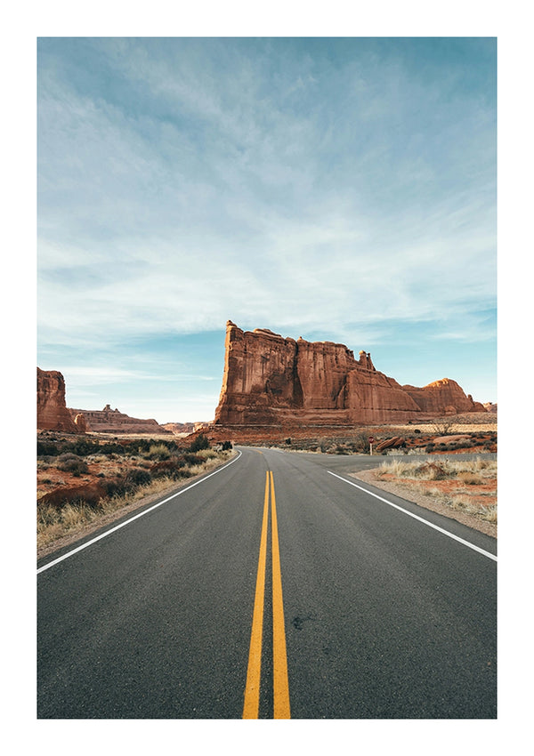 Een rechte weg met dubbele gele lijnen leidt naar grote, rode rotsformaties onder een overwegend heldere hemel met weinig verspreide wolken. Woestijnvegetatie omlijnt de zijkanten van de weg en de rotsformaties verschijnen op de achtergrond, waardoor een scène wordt vastgelegd die perfect is voor een CollageDepot Desert Road Schilderij.