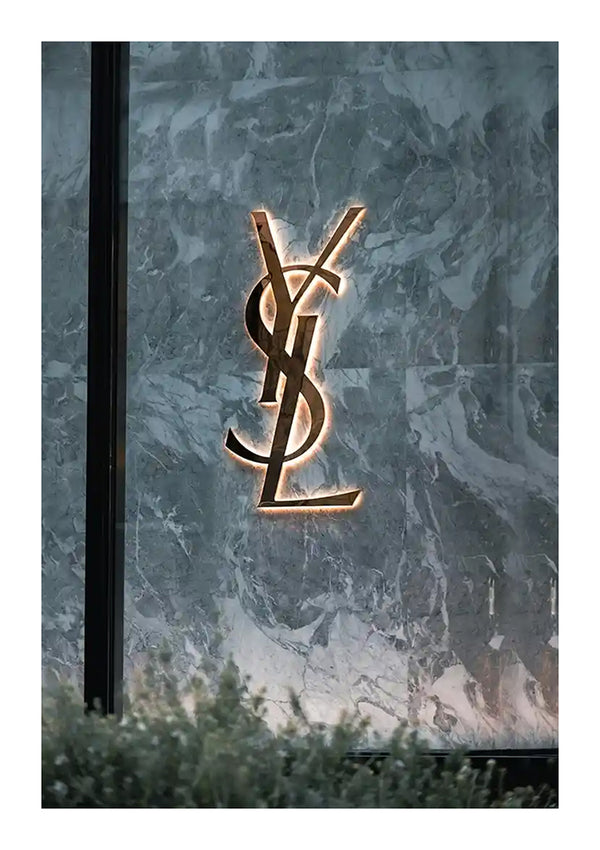 Op een grijsmarmeren muur is een gouden YSL-logo aangebracht, met subtiele achtergrondverlichting die de contouren van de letters benadrukt. Het YSL Logo Schilderij van CollageDepot is gemaakt als een elegante wanddecoratie en toont groen dat gedeeltelijk zichtbaar is onderaan de afbeelding.-