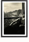 Zwart-witfoto van een persoon in een donkere jurk, liggend op een balkonleuning met een stedelijke dakachtergrond. De scène heeft een vintage uitstraling, perfect voor stijlvolle wanddecoratie met het Balkonzicht schilderij van CollageDepot, en het gezicht van de persoon is niet duidelijk zichtbaar.