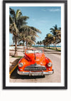 Een rode vintage auto staat geparkeerd in een met palmbomen omzoomde straat vlakbij een strand met een helderblauwe lucht op de achtergrond. De auto, die doet denken aan een oldtimerschilderij, staat naar voren gericht met het kenteken in zicht. Palmbomen en de oceaan zijn zichtbaar, waardoor een tropische setting ontstaat, perfect voor wanddecoratie met een magnetisch ophangsysteem. Dit idyllische tafereel zou prachtig worden vastgelegd door het Zomerse Vibes Oldtimer Schilderij van CollageDepot.