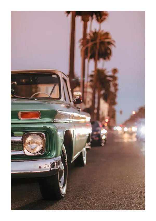 Een groene cbb 004 - vintage van CollageDepot staat tijdens zonsondergang geparkeerd in een straat vol palmbomen. De koplampen van de vrachtwagen en andere auto's op de achtergrond branden en in de verte zijn onder een schemerige hemel gebouwen zichtbaar.-
